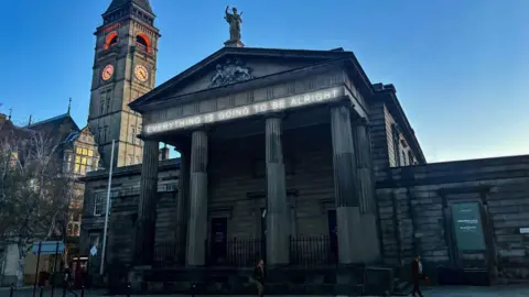 A white neon sign reading "everything is going to be alright" is installed on a court building with four pillars.