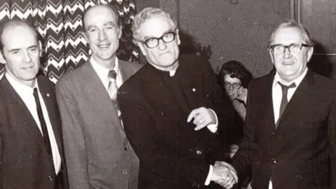 Jim McQuaid Fr Austin Eustace in a black and white photo with three other man and a woman in the back ground. Fr Austin is wearing his black suit with priest collar. He has white/grey curly short hair and is wearing black-framed glasses. He is shaking the hand of a man to his left but looking directly at the camera. 