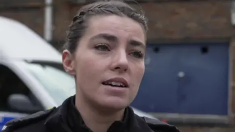 PC Jess Surman gives a TV interview outdoors. Her dark hair is tied back in braids and she wears a police uniform.