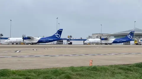 Two Blue Island planes parked up on the tarmac at an airport. The terminal building is in the background of the shot.