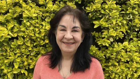 Cheryl Kenealy is a woman with dark hair and dark eyebrows, wearing a pink jumper. She is standing in front of a green bush and smiling into the camera.