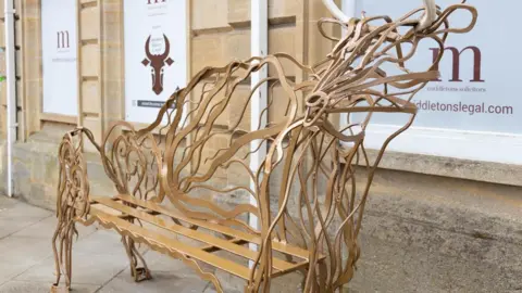 Bertie Coy A bench in the shape of the prehistoric aurochs sitting outside a building clad in Cotswold stone. The animal looks similar to a bull, with broad shoulders and horns. The bench is metal and the sculpture is made up of thin waved lines.