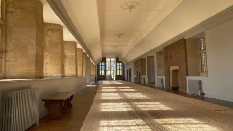 National Trust/Pippa Gibson A long room with stone windows to the left letting in lots of light. There is a glass door at the end of the room with more sunshine flowing through it. A long rug runs the length of the room, which has a fireplace and modern radiators along its walls. 