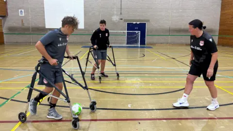 A female football coach works with two teenage boys using mobility frames as they play football in an indoor sports hall.