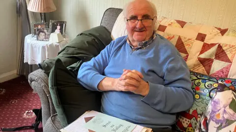 An elderly man who is partially bald with some short white hair and is wearing a blue sweater over a checked shirt. He is wearing glasses and has a pile of documents sitting on his lap. Behind him is a small table with a number of framed pictures on it.