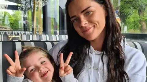 A woman and her daughter are sitting on a bench. The little girl is holding her hands up in "V" signs and the woman, who is wearing a white hooded top, is smiling.
