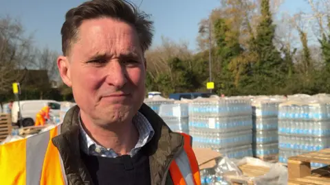 Charlie Maynard has short brown hair and is wearing an orange hi-vis jacket over a brown coat. He is standing in front of large pallets full of bottled water.