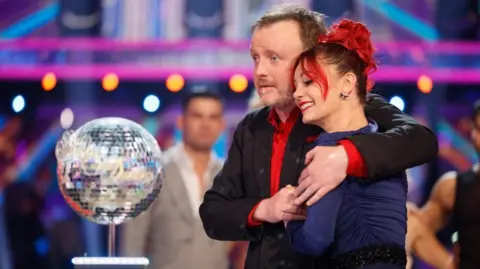 BBC/PA Chris McCausland stands wearing a red shirt and suit jacket with his arms around a woman with red hair and wearing a long-sleeved dress. A disco ball statue is on a stand in front of them. 