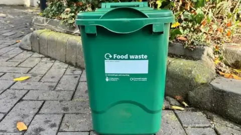 A green food waste collection bin from Cornwall Council in a paved area outside a house. The bin is in front of a bush.