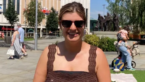 Hannah smiles for the camera. She is wearing a brown dress and sunglasses.
