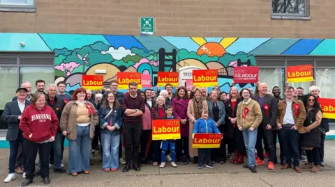 A group of people standing in front of a mural. They have red rosettes on their clothes and are holding red and yellow cards with the words vote Labour written on them.