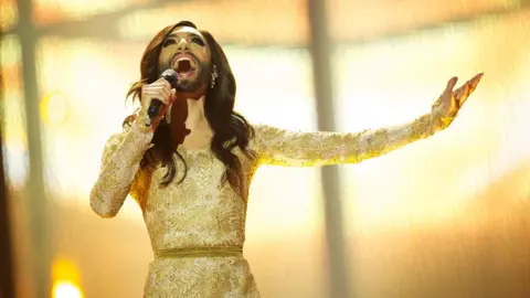Getty Images bearded drag queen conchita wurst performing at the 2014 eurovision song contest. she is wearing a gold brocade dress, has long dark hair and is holding a microphone while looking upward
