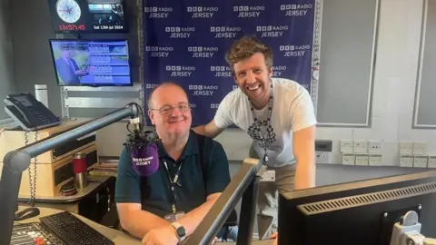 Phil Taylor, a man with short brown hair and glasses, wearing a dark blue polo shirt. He is sitting in a BBC Radio Jersey studio, in front of a purple microphone. TV screens are visible behind him, to the left of the image. To the right of the image, Warren Muggleton, a man with short brown-blonde hair, is standing with his arm on Phil's shoulder. Warren is wearing a white t-shirt and chinos. Both have BBC lanyards around their necks, and both are smiling directly at the camera.