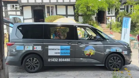 A gun-metal grey people carrier with sticker and sign writing is side on in front of a tudor style black and white building.