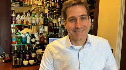 The image shows a man standing behind a bar counter. He is wearing a light blue, long‑sleeved, button‑up shirt and is positioned slightly to the right side of the frame.