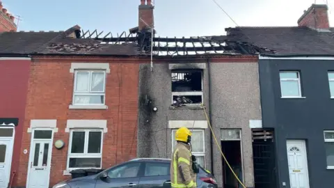 A residential street where a fire has ripped through the roof of three properties. A firefighter is in front of the houses and a grey car is also parked there.