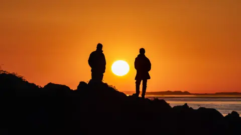 Robert Westerman Two silhouetted figures stand on rocks against a vibrant sunset, casting a warm glow over the serene ocean.