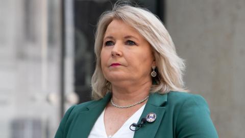 Pat Cullen standing outside. She's wearing a green jacket and white top, and silver earrings and a necklace. She has shoulder length blond hair