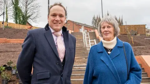 A man in a long navy overcoat with a pink tie and shirt stands next to an elderly woman wearing a blue coat and black gloves. The pair stand in front of a walkway with dirt on either side of it which has plant pots and plants across it.