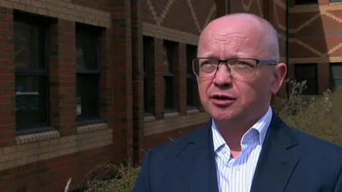 A man in glasses and a white striped shirt and navy blazer stands by a brick building looking off camera. 