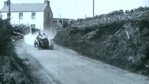 Manx National Heritage A black and white image of an early motorcar driving down a road lined with tall verges and a stone wall on the right. There is a white house behind at the side of the road and smoke coming from the back of the car.