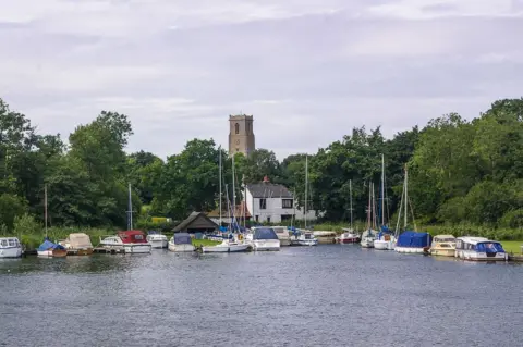 Ian Capper/Geograph Church tower in distance. In foreground is an expanse of river water with about 20 boats of different styles, including sailing boats and mini cruisers moored outside a building which looks like a pub. 