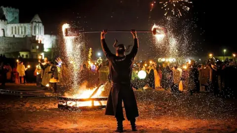 Corraste Photography A man in a horned helmet and cloak on Peel Beach holding aloft a rod that is lit at both ends as part of a fire display in front of a bonfire. Peel Castle is illuminated in the background.