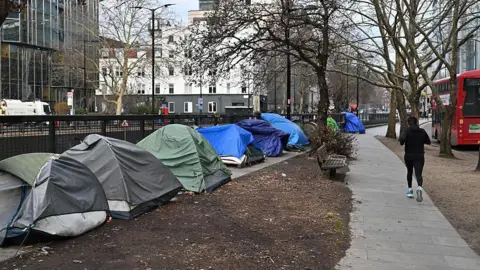 Getty Images The side of a road is lined with tents and person runs past them on the path. A London red bus is seen going by on the right hand side of the runner.
