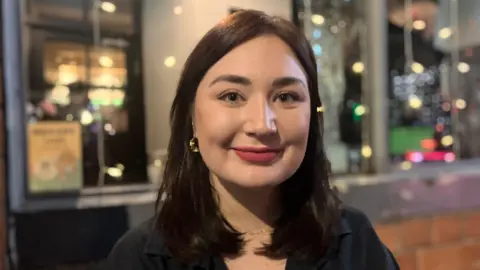 A woman with dark shoulder length hair and blue eyes, wearing gold loop earrings and a black shirt. She is standing in front of a building with sparkling lights in the windows.