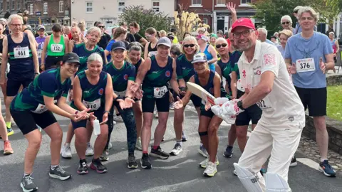 A man wearing cricket whites is standing in the middle of the photo. He is wearing gloves, shin pads, and is holding a cricket bat as if to strike the ball. He has glasses and a red baseball cap on. He is surrounded by runners pretending to be ready to catch the ball.