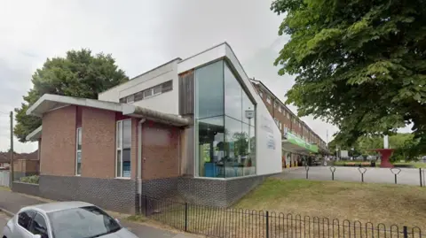 Google A building behind a fence with a silver car towards the front of the image
