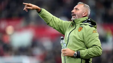 PA Media Hull City manager Tim Walter gesticulating form the touchline wearing a lime green jacket and holding a bottle of water