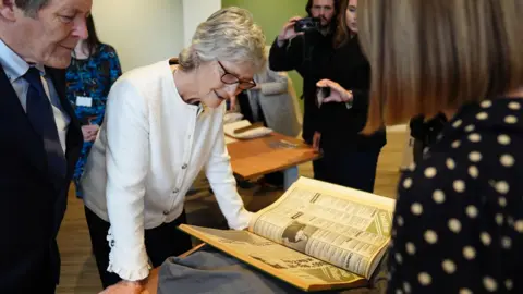 PA Media Irish president Catherine Connolly during a visit to Leeds University, as part of her official visit to the UK. She has short white hair wearing glasses and a cream cardigan. She is leaning over a table looking at a large open book. Other people are around her.