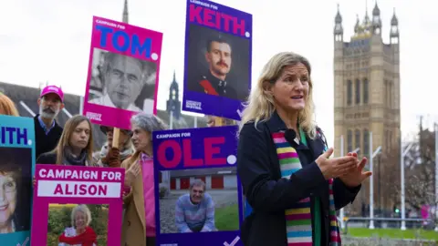 Labour MP for Spen Valley Kim Leadbetter wears a colourful stripey scarf while gesturing and speaking in front of parliament with campaigners in the background holding placards showing faces of people affected by the current law against assisted dying, in London in December.