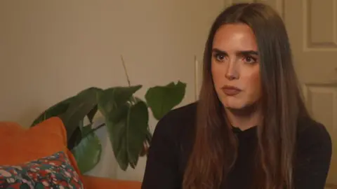 Hannah Strickland is sitting on a large orange sofa with a plant in the background