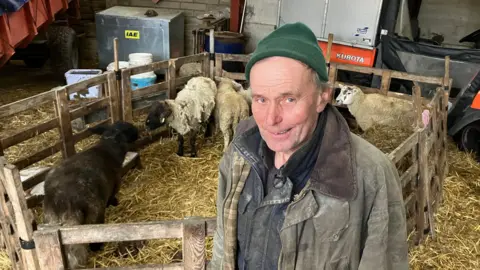 Andy Gill/BBC Alan Jackson, 65, who is wearing a green woollen hat and a green overcoat, stands in front of a small wooden pen containing five sheep with farm equipment in the background. 