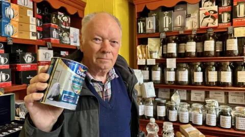 John is holding a can of paint to the camera. There are lots of bottles with loose tea in the on shelves in the background.