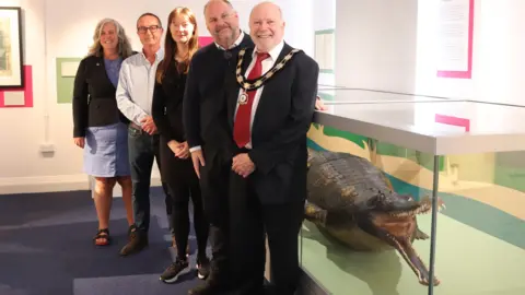 Swindon Borough Council Heidi Alexanderm, Andrew Cross, Frances Yeo, Councillor Jim Robbins, and Councillor Neil Hopkins standing in front of the glass cabinet where the gharial is displayed.