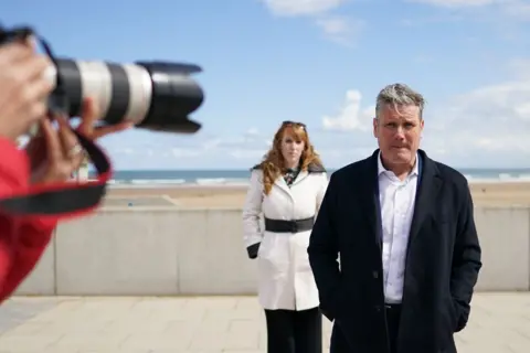 Getty Images Labour Party leader Sir Keir Starmer and Angela Rayner, deputy leader and chairwoman of the Labour Party visit Seaton Carew seafront on 1 May 2021 in Seaton Carew. Sir Keir is in the foreground with his hands in his pockets, with Rayner behind him. A camera shoots a photo, with the beach behind them.