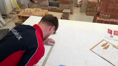 A man with short brown hair leans over a drawing of brickwork on a large white sheet of paper. He is measuring with a ruler over the drawing. A clipboard with other drawings is on the right, with piles of bricks behind. 