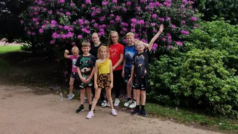 Nikki Blackham Seven children of different ages stand together by a large floral bush outside. They are wearing different coloured shorts and T'shirts and trainers.