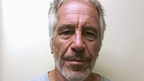 Mughshot of Jeffrey Epstein, who has grey hair, a beard, and is wearing a grey t-shirt