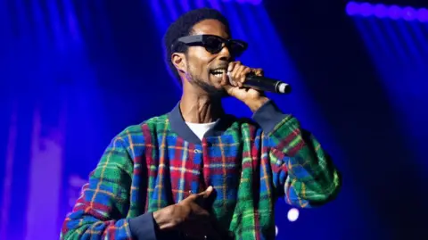 Getty Images D Double E performs on day 1 of Reading Festival 2023. he is holding a black microphone, wearing dark sunglasses, and has a green, blue and red striped sweater top on. The background is of dark blue coloured lights.