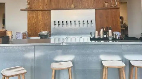 Liz Cottam A bar area at a restaurant with a chrome-coloured counter, topped with drinking measures. In front are four wooden bar stools. Behind the counter is a wood-panelled wall with a chrome panel in the centre. Taps are mounted on the panel.
