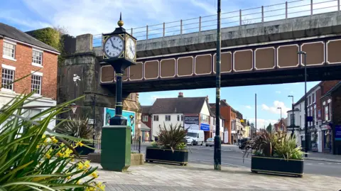 A town centre street with a clock on a pillar, a railway bridge and a road junction