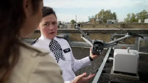 A female police officer in uniform holds up a drone and gestures towards it as she explains to a reporter how it works. They are stood on a roof top