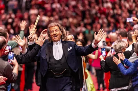 Marcel van Hoorn/EPA-EFE Pemain biola Andre Rieu, mengenakan setelan hitam dengan kemeja putih, berjalan, tersenyum, tangan terbuka lebar, melewati kerumunan orang, beberapa mengambil foto dengan ponsel, di Maastricht, Belanda.