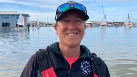 Nicky Will is stood in front of the water smiling straight at the camera. She is wearing a life jacket, a branded Guernsey Rowing Club fleece and a hat with sun glasses.