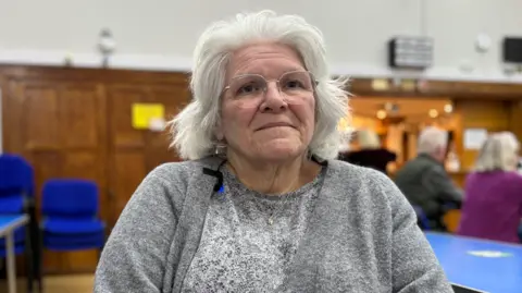 Charlie O'Loughlin A woman sits in a large wood-panelled hall and looks towards the camera. She has white hair framing her face and wears a grey cardigan and grey T-shirt. She has thin wire-framed glasses. Behind her, the background is blurred, but the backs of people sitting at tables can be seen, along with stacks of blue chairs.