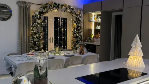 IWC Media A Christmas themed kitchen. A large archway made of foliage, baubles and warm yellow Christmas lights borders the patio doors. A dining table, covered in fairy lights and a star, looks set for Christmas dinner.